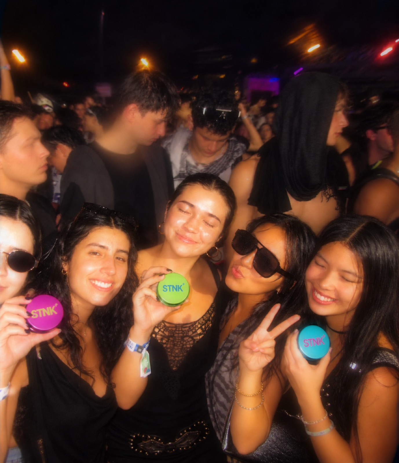Five friends at a night event, three holding up STNK tins in magenta, lime and teal